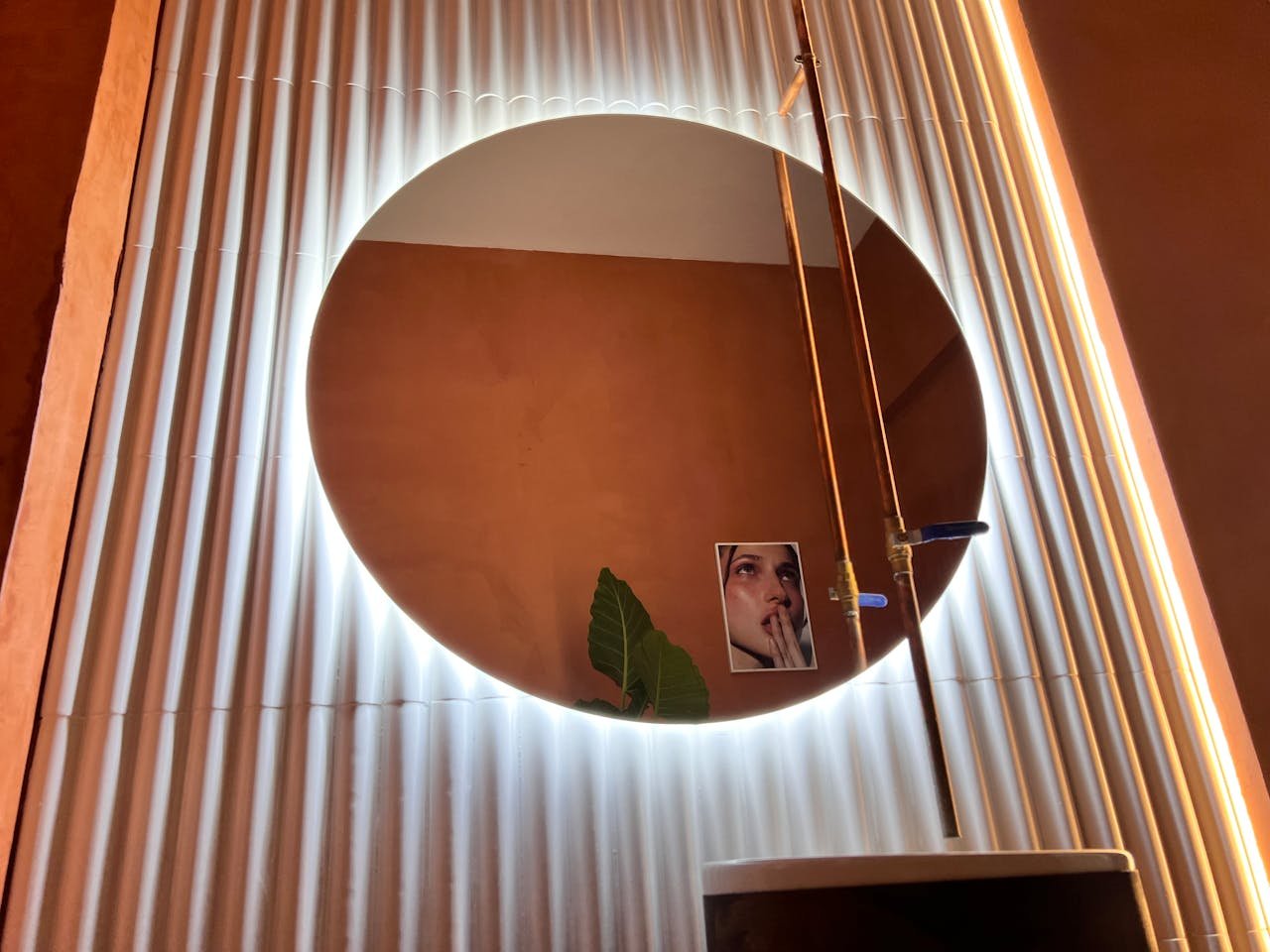 gallery-3 A modern circular mirror with LED lights on a paneled wall, reflecting art and foliage.